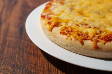Cooked cheddar cheese pizza on a white plate and dark brown wooden table. Italian style dish. Fast food industry product. Close up, selective focus