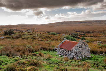 Fototapeta na wymiar Small stone building with rusty red metal roof with beautiful nature scenery in the background. Connemara area, county Galway, Ireland. Storage hut in a field. Historical building. Cloudy sky