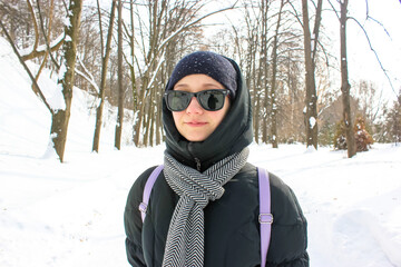 Fototapeta na wymiar A young smiling woman on a walk in a winter park. A white girl standing outside in a warm down jacket, sunglasses, with scarf around her neck walking outside in a snow-covered forest. Female traveler.