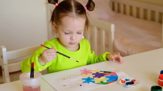 A little sweet girl dips a brush into a glass of water and carefully paints her picture with gouache. A child learns to draw. A child draws a multicolored heart from puzzles