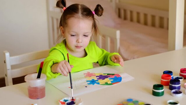 A two-year-old child draws a multicolored heart from different pieces of puzzles with watercolor paints. The girl likes to make a postcard for a friend with autism syndrome
