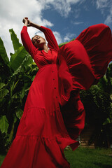 Beautiful young woman in flowing red dress posing front of tropical green leaves. Fashion portrait of attractive model girl at nature background. Selective focus.