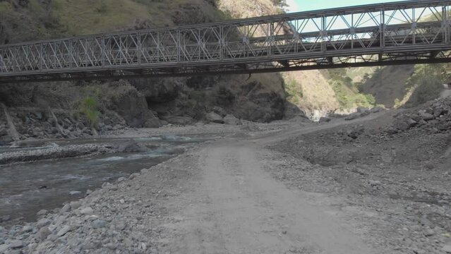 metallic bridge spanning over rocky river bed in mountainous valley surrounded by mountains trees bushes brush rocks dust in Kabayan Benguet Philippines fast low flying aerieal approach