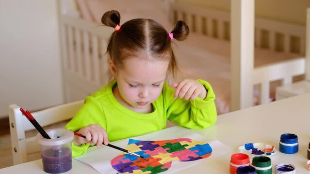 A girl with autism syndrome paints over a drawing with a picture of a heart from puzzles with a brush.