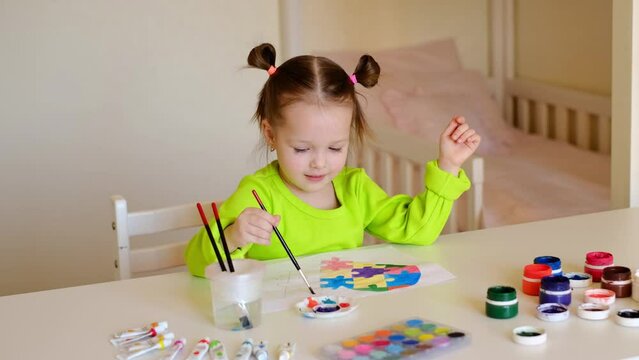 A preschool-age girl diligently paints a drawing with a picture of a heart from puzzle pieces. Colorful picture for Autism Day