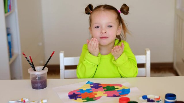 A funny little sweet girl sends blows kisses to the whole world. A child is sitting at a table with a drawing for friends suffering from autism syndrome on the eve of Autism Awareness Day. 