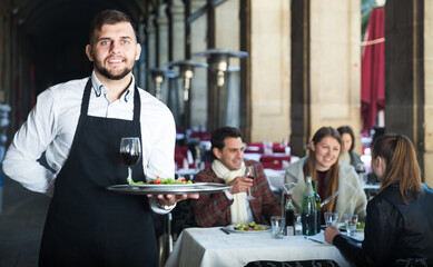 Smiling waiter holding tray at restaurant with customers his behind