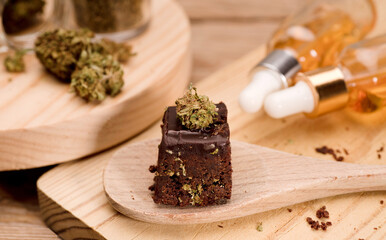 Fototapeta premium Chocolate cupcake muffins with cannabis buds in wooden spoon, oil pot and cooking jars. Marijuana hemp in food dessert. Chocolate herb muffins cooking. Marijuana cake on a brown wooden table.