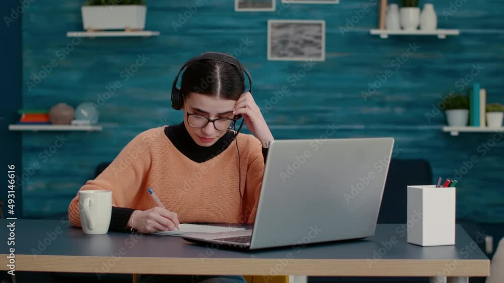 Poster woman with headphones talking on online video call and taking lecture notes on notebook, learning in