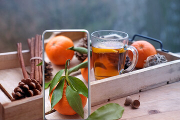modern mobile phone with screen, glass of hot tea on tray, delicious vitamin tangerines with leaves, cinnamon sticks, pine cones, cozy mood, concept vacation, weekend in nature, breakfast on terrace