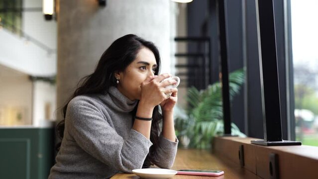 Young woman sitting in a modern Coffeehouse café drinking a cup of hot tea coffee while checking his smartphone device sharing post on social media account