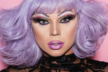 portrait of man with bright makeup, wearing purple wig and looking at camera on pink.