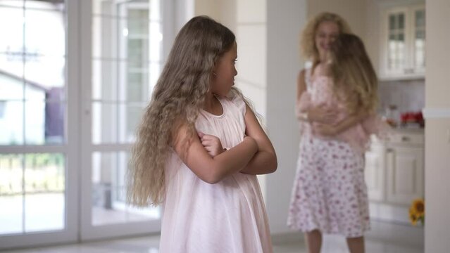 Envious sad girl with crossed hands standing in living room looking back at blurred mother having fun with little sister. Portrait of upset Caucasian child standing at home indoors thinking