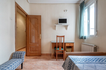 Bedroom with bed and footstool, desk with chair, white tv, matching wooden chair, plastic top, white aluminum radiator and window with green curtain