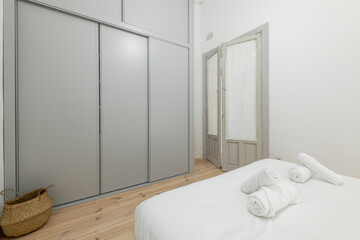 Fototapeta na wymiar Bedroom with white bedding, rolled towels on the beds, gray sliding-door wardrobe and wooden door with pinewood floorboards