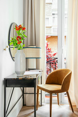 Steel desk with wooden top, large gray vase with flowers, circular mirror and chair upholstered in orange velvet and balcony with curtains and net curtains