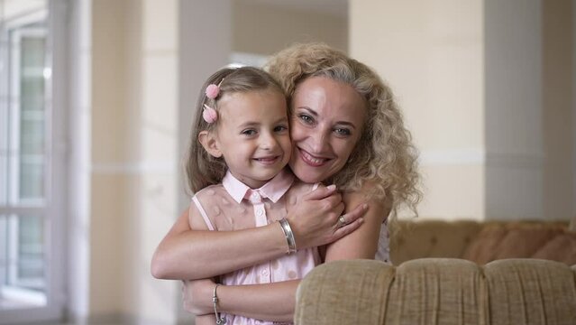 Portrait of happy little girl smiling looking at camera as woman hugging child posing in slow motion. Front view medium shot of Caucasian daughter and mother at home in living room indoors