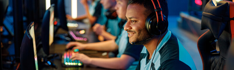 Happy cybersport gamer playing online game while sitting with other team members in front of PC monitors in computer club © Yaroslav Astakhov