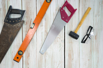 The equipment for woodworking is organized neatly on the wood floor. Ordered as follows, old saw full of rust, orange magnetic aluminum level, red hand saw, yellow mallet, black dustproof glasses.