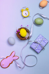 Festive Easter composition - yellow egg in bird's nest, Easter bunny, envelope letter, retro alarm clock, flowers on light lilac background top view. Vertical holiday wallpaper flatly. Easter time.