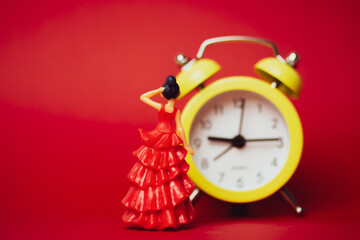 Toy woman in red elegant dress fixing hair looking at yellow alarm clock. Girl getting ready for date concept. Time to sign up for flamenco dance studio. Punctual woman concept. Cinderella. Copy space