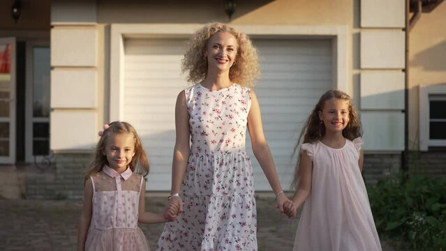 Dolly shot of confident Caucasian mother and daughters holding hands walking in slow motion outdoors. Front view of woman and girls strolling on front yard at sunset smiling