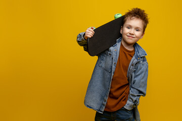 Cute little boy 6-7 years old, in stylish clothes, holding a skateboard in his hand, isolated on a yellow background, a children's studio portrait. the concept of a child's lifestyle, sincere emotions
