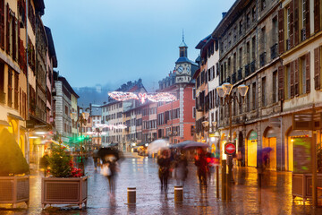 View of Place Saint-Leger, main pedestrian street in historic center of French city of Chambery decorated with traditional Christmas lights overlooking clock tower on rainy winter evening.
