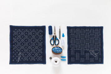 Process of embroidering two geometric patterns with white threads on blue fabric in traditional Japanese Sashiko style. Accessories for embroidery on white background. Copy space, flat lay, close-up
