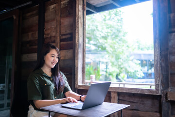 Happy of asian freelance people business female  wearing brownish green dress stylish hipster casual working with laptop computer with coffee cup beside the window of a wooden house background