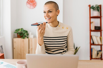 Smiling young adult woman using mobile phone while sitting at home office working on laptop computer