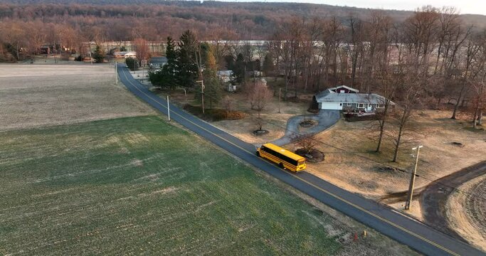 School bus drives on road through rural American countryside. Homes at winter sunrise. Aerial view.