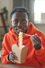 Vertical medium close-up shot of modern African American young man sitting at table at home eating...