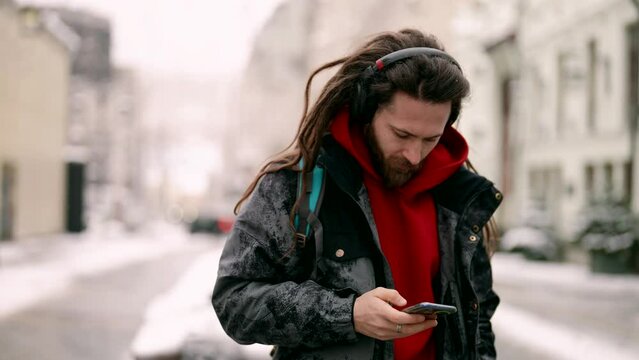 trendy dressed young man with dreadlocks is walking in city and listening to music by wireless headphones