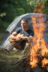 Romantic guy camping outdoors and sitting near tent. Handsome man playing guitar in forest with bonfire. Country music song. Guy by a fire in the forest, weekend near campfire.