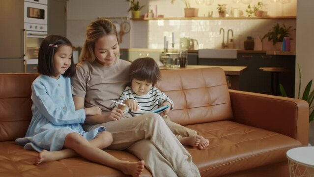 Loving Asian Mom and Little Daughter and Son Sit Together on Sofa at Home With Tablet Device Draw With Pencel, Using Education Application for Easy and Funny Learning, Modern Tech and Generation