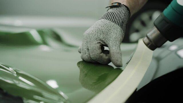 Man vinyl wrapping the car, smoothing the wrap with his hands and construction dryer. Process of vinyl wrapping a car in a garage. Khaki green vinyl car wrap. High quality 4k footage
