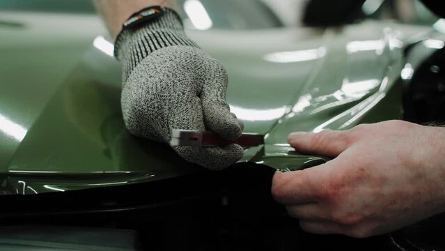 A specialist cuts khaki green vinyl car wrap on a car hood. Close up shot of vinyl wrapping a car hood. Process of wrapping a car in a garage, using a blade to trim vinyl film. High quality 4k footage