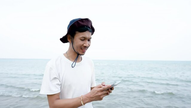 4K 50fps, Good-looking Asian man, standing by the sea on the phone playing in the evening with clear air, cool breeze, happy smiling face.