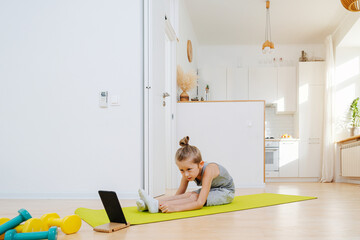 Cute boy doing yoga in front of a phone in a holder, following instructions to the best of his...