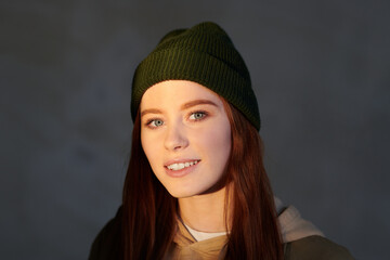 Horizonatal close-up shot of attractive young woman with darl red hair wearing cap smiling at camera, gray background