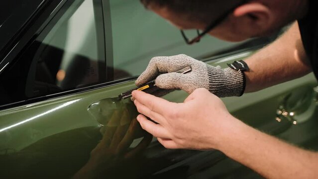Close up shot of cutting a vinyl car wrap on a door. A car wrapping specialist cuts dark green vinyl film. Process of vinyl wrapping a car in a car studio. High quality 4k footage