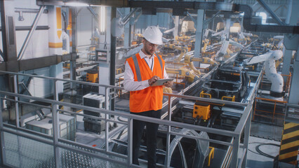 Male employee in uniform and hardhat browsing tablet against automatic machines on modern vehicle...