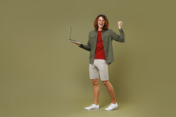 Full size body length fun young brunet curly man 20s wears khaki shirt hold use work on laptop pc computer doing winner gesture clenching fist isolated on plain olive green background studio portrait.