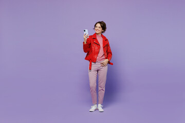 Full body young smiling happy woman 20s wear red leather jacket hold in hand use mobile cell phone isolated on plain pastel light purple background studio portrait. People lifestyle fashion concept.