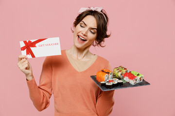 Young cheerful woman in casual clothes hold in hand makizushi sushi roll served on black plate traditional japanese food gift certificate coupon voucher card isolated on plain pastel pink background