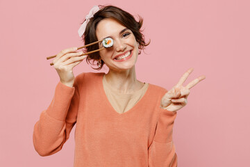 Young cheerful smiling happy fun cool woman in casual clothes hold in hand cover eye with makizushi sushi roll traditional japanese food showing victory sign isolated on plain pastel pink background