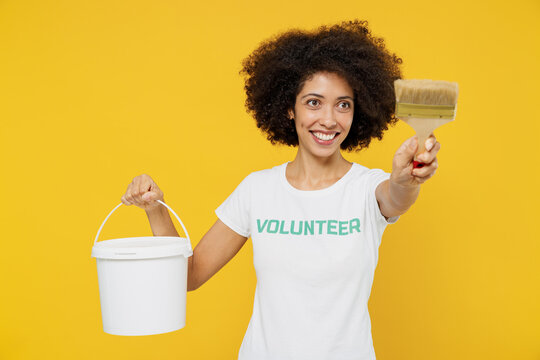 Young fun woman of African American ethnicity in white volunteer t-shirt hold paint bucket brush working isolated on plain yellow background. Voluntary free work assistance help charity grace concept.