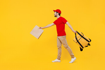 Full size delivery guy employee man in red cap T-shirt uniform work as dealer courier hold brown blank craft paper takeaway bag mock up thermal food bag backpack go isolated on plain yellow background