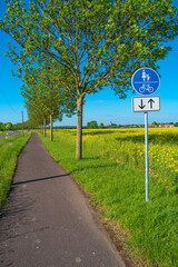 Beautiful farm landscape with rapeseed blossom field and a cycling and hiking lane or path in Germany, Spring, at sunny day and blue sky, with a signpost for bicycles and people.
